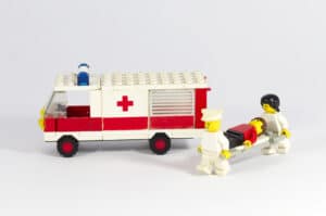 Ein LEGO-Krankenwagen mit dem Symbol des Roten Kreuzes steht neben einem Krankenwagen. Zwei LEGO-Figuren, als Sanitäter verkleidet, tragen eine weitere Figur auf einer Trage.