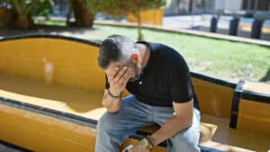 Ein Mann in schwarzem Hemd und Jeans sitzt draußen auf einer gelben Bank und bedeckt sein Gesicht mit einer Hand. Er scheint verzweifelt oder in tiefe Gedanken versunken zu sein. Der Hintergrund besteht aus Grün und verschwommenen Außenelementen.