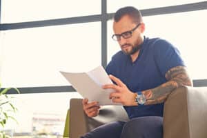 Ein Mann mit Brille, Bart und Tattoos auf dem Arm sitzt in einem Sessel neben einem Fenster und liest konzentriert Zeitung. Er trägt ein blaues Hemd und eine Armbanduhr. Helles Tageslicht erhellt den Raum.